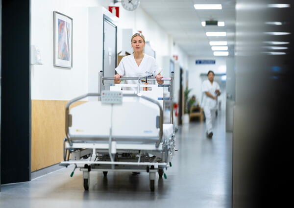 En sykepleier i uniform triller en sykeseng i en korridor.