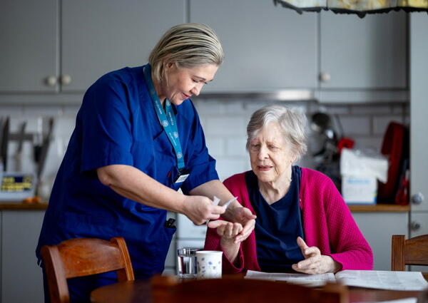 En hjemmesykepleier i blå uniform legger medisiner i hånden på en eldre kvinne som sitter på en kjøkkenstol.