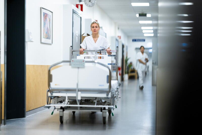 En sykepleier i uniform triller en sykeseng i en korridor.