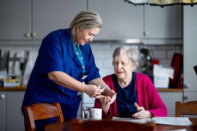 En hjemmesykepleier i blå uniform legger medisiner i hånden på en eldre kvinne som sitter på en kjøkkenstol.