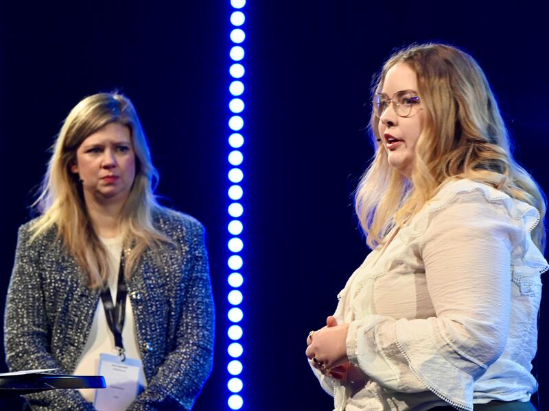 To kvinner i samtale på en scene. Den ene ser på den andre, som ser ut mot publikum.