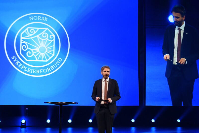 En mann med dress og slips står på en scene med logoen til Norsk Sykepleierforbund bak seg.