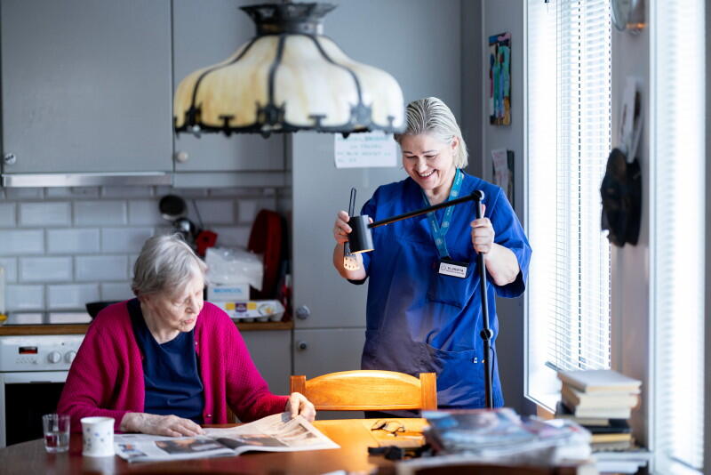 En dame i blå uniform står på kjøkkenet til en eldre dame som sitter på en kjøkkenstol og leser avisen.