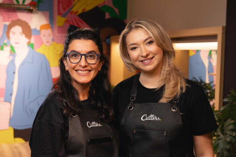 To kvinner med forkle som det står "Cutters" på, smiler til kamera.