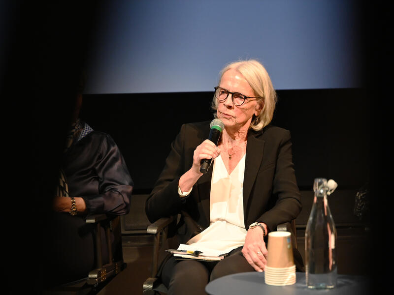 Kvinne med briller snakker i en mikrofon og ser mot venstre.