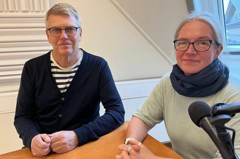 En mann og en kvinne, begge med briller, sitter ved et bord og ser mot kameralinsen.